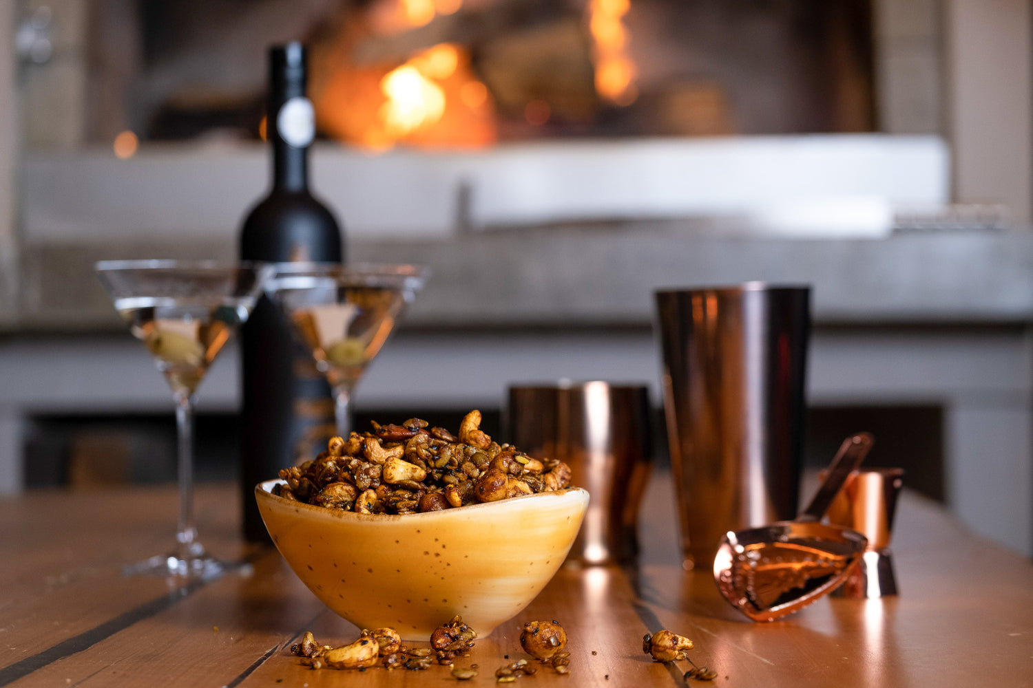 A bowl of Umami Larder Silk Road Seeds and Nuts sits on a table with a cocktail shaker and two martini glasses sitting in the background. A fire burns beyond that.