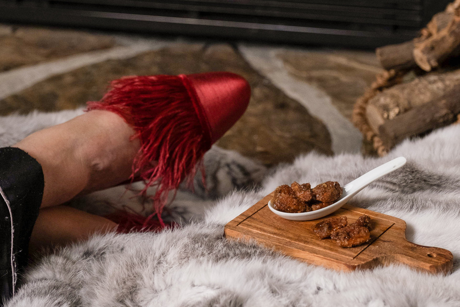 A women in slippers sits by the fire with a small wooden board of Umami Larder Spiced Bourbon Pecans beside her. 